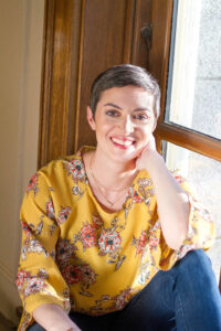 Woman in yellow shirt sitting in window, smiling at camera